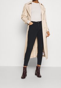 Gabardina beige con cintura ceñida sobre un suéter claro, combinada con jeans skinny oscuros y botas de tacón tobilleras burdeos.