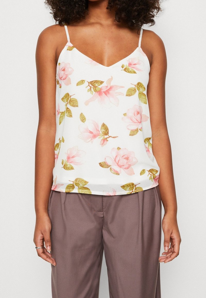 Femme portant un haut blanc sans manches avec des motifs floraux roses et un pantalon marron ample, debout devant un fond uni.