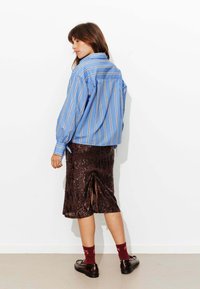 Chemise rayée bleu et blanc à manches longues, associée à une jupe midi marron à paillettes avec une fente latérale. Chaussures foncées et chaussettes bordeaux.