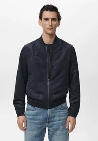 Chaqueta bomber de ante azul marino con puños y bajo de canalé, cierre frontal de cremallera y hombros sin costuras, combinada con jeans de color azul claro.