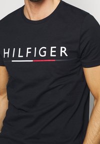 Czarna bawełniana koszulka z klasycznym okrągłym dekoltem i krótkimi rękawami, z centralnie umieszczonym białym logo "HILFIGER" oraz czerwonymi i niebieskimi poziomymi akcentami.