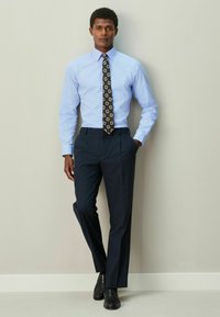 Mænds business outfit med en lyseblå skjorte, mønstret slips med firkanter og mørke bukser med nålestriber. Sorte lædersko.