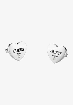 Orecchini a forma di cuore in argento con logo in rilievo "GUESS EST.1981" e un piccolo accento di pietra trasparente. Texture metallica liscia.