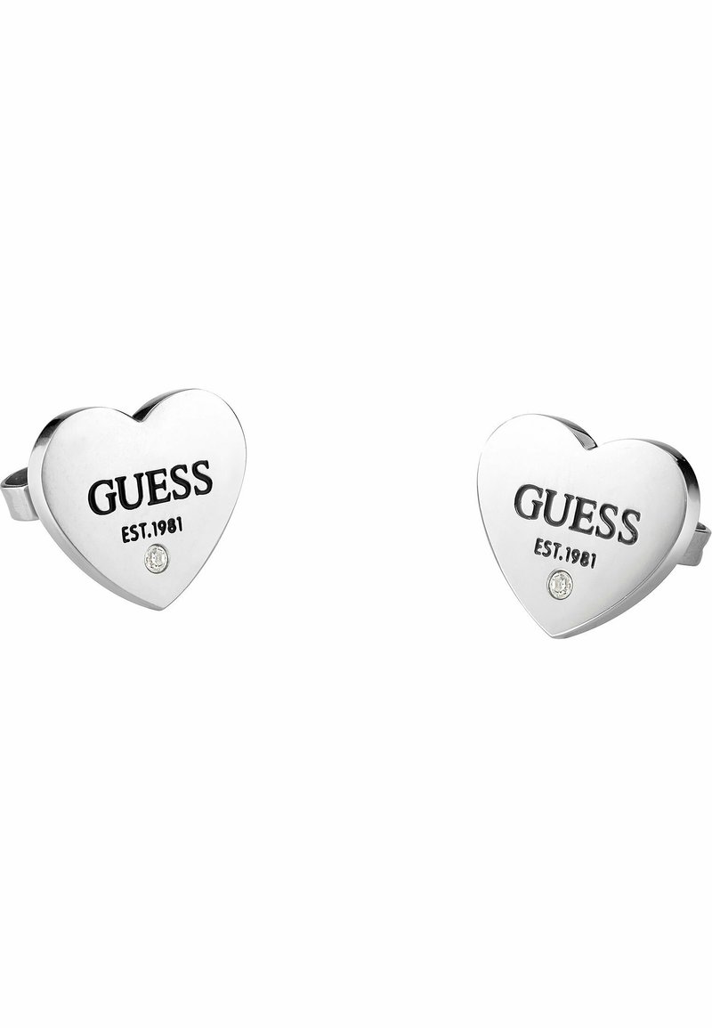 Boucles d'oreilles en argent en forme de cœur, arborant le logo en relief "GUESS EST.1981" et une petite accent de pierre précieuse transparente. Texture métallique lisse.