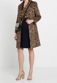 Manteau en imprimé léopard marron et noir, associé à une jupe noire et des sandales dorées. Présente un design structuré et des manches longues.