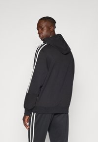 Molete preto com riscas laterais brancas, textura suave e capuz. Modelo está a usar calças de treino pretas combinando, em pé num fundo neutro.