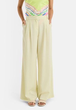 Mujer con pantalones anchos color beige claro con pliegues delanteros, manos en los bolsillos, combinados con sandalias abiertas color nude y una blusa colorida.