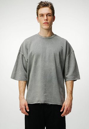 Junger Mann mit kurzen Haaren, der ein übergroßes graues kurzärmeliges Hemd und schwarze Hose trägt, steht vor einem schlichten weißen Hintergrund.