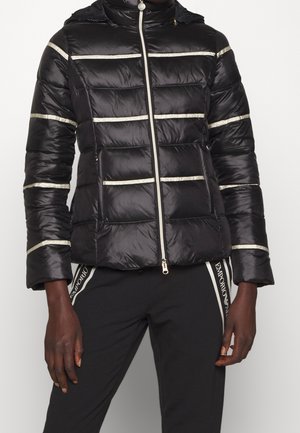 Doudoune noire avec des rayures horizontales argentées et une fermeture éclair à l'avant, portée avec un pantalon noir avec des bandes latérales blanches brandées.