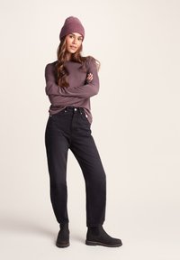 Long-sleeve purple shirt, black high-waisted jeans, and black ankle boots. The model wears a ribbed purple beanie, arms crossed.