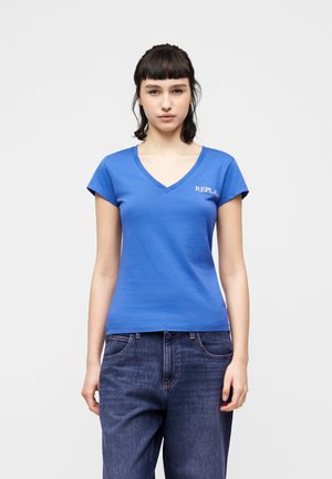 Młoda kobieta o ciemnych włosach, ubrana w niebieską koszulkę z dekoltem w serek z logo „REPLAY” oraz luźne ciemnoniebieskie dżinsy, stoi na tle jednolitego jasnego tła.