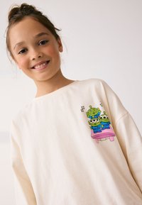 Cream-colored sweatshirt featuring a graphic of three green aliens sitting on a pink bench, with stars above them. Soft fabric and relaxed fit.