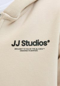 Beżowa bluza z kapturem na zamek o miękkiej teksturze. Posiada czarne logo "JJ Studios" oraz dodatkowy tekst nadrukowany poniżej w czytelnej czcionce.