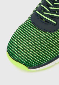 Partea superioară a pantofului sport, cu dungi verzi și negre, confecționată dintr-un material texturat, cu vârf rotund și talpă neon verde contrastantă. Dispune de șireturi negre și ochiuri pentru șireturi.