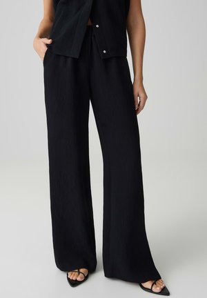 Vrouw draagt zwarte broek met wijde pijpen en een zwart mouwloos topje, staat met één hand in de zak, draagt zwarte open-teenhakken.