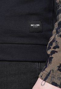 Granatowa bluza z prążkowanym dołem, z czarną wszywką. Noszona z ciemnymi dżinsami. Rękaw ma wzór przypominający kamuflaż w brązowo-czarnych kolorach.