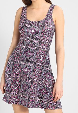 Femme portant une robe sans manches, longueur aux genoux, avec un motif géométrique tribal aux couleurs violet, rose, noir et blanc.