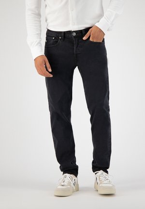 Mann trägt eng anliegende schwarze Jeans, weißes Langarmshirt und weiße Sneakers mit schwarzem "V"-Logo, steht mit einer Hand in der Hosentasche.