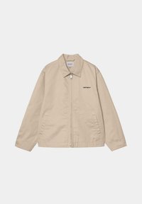 Chaqueta beige con cremallera frontal, cuello puntiagudo y dos bolsillos laterales. Presenta un pequeño logo negro en el lado izquierdo. Textura de tela suave.