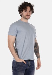 Maglietta a maniche corte blu chiaro in morbido tessuto con scollo rotondo, indossata da un modello con capelli ricci e tatuaggi, abbinata a jeans scuri.