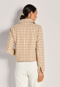 Vrouw met donker haar in een paardenstaart, gekleed in een beige en witte houndstooth jasje en witte broek, staand tegen een simpele lichte achtergrond.
