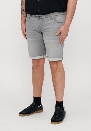 Hombre vistiendo pantalones cortos de mezclilla gris claro enrollados, calcetines negros, zapatillas negras y una camiseta negra, de pie contra un fondo liso.