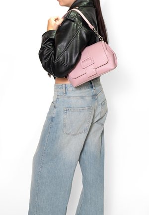 Donna con giacca di pelle nera e jeans azzurri chiari che porta una borsa a tracolla in pelle rosa su sfondo bianco.