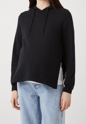 Personne portant un sweat à capuche noir avec cordons à l'avant et un jean en denim bleu clair sur un fond blanc uni.