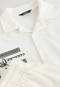 Chemise blanche texturée à manches courtes boutonnée avec col et short à taille élastique, présentant un détail noir avec motif géométrique et feuille.