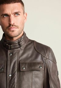 Braune Lederjacke mit hohem Kragen, zwei Fronttaschen mit Druckknöpfen und gesteppten Akzenten an den Schultern. Glatte Textur und taillierte Passform.