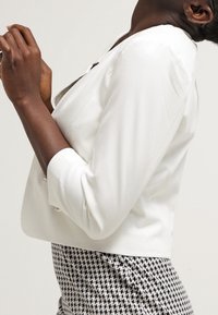 Blazer blanc à coupe ajustée, fermeture à deux boutons, et manches trois-quarts, associé à une jupe à motif pied-de-poule noir et blanc.