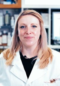Une personne en blouse de laboratoire blanche portant un haut noir, les cheveux blond clair et un microphone ; des étagères avec divers produits sont floues en arrière-plan.