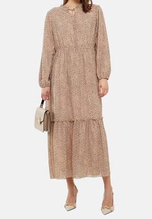Mujer con un vestido largo beige con puntos negros, sosteniendo un bolso beige y llevando tacones puntiagudos beige con detalle de cadena dorada.