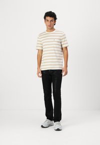 Striped short-sleeve t-shirt in beige and white, featuring a chest pocket and logo. Paired with black pants and gray sneakers.