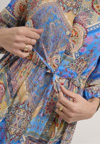 Farverig mønstrede kjole med blå, rød og gylden nuancer. Stoffet har komplekse paisley-designs, knapper og en bundet taljedetalje.