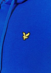 Sininen kangas, jossa vasemmalla puolella pieni keltainen kirjailtu lintulogo ja vastaavansininen nyörikoriste.