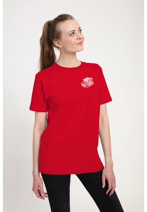 Kvinde med langt brunt hår sat op i en hestehale iført en rød t-shirt med et lille lyserødt læbeaftryk på brystet, der kigger opad og smiler.