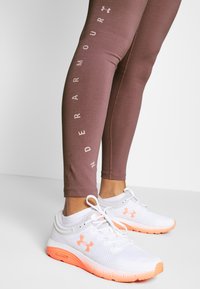 Brązowe legginsy z białym logo "Under Armour" wzdłuż boku, zestawione z białymi i pomarańczowymi butami do biegania, wyposażonymi w teksturowaną siateczkę i amortyzowaną podeszwę.