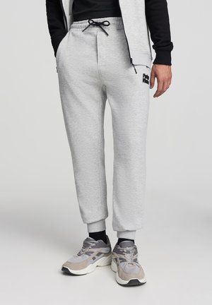 Pantaloni jogger grigio chiaro con vita elasticizzata, cordino, tasche laterali e polsini a costine. Abbinati a scarpe sportive grigie e beige.