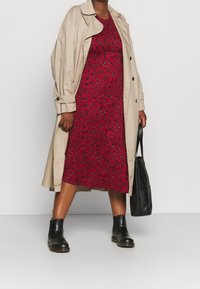 Trench-coat beige porté sur une robe rouge à motif floral noir et vert, associé à des bottines noires et un sac à main noir.