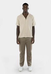 Kremfarget kortermet skjorte med knapper og teksturert finish, kombinert med beige bukser og hvite joggesko. Enkel og avslappet passform.