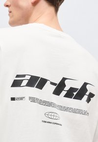 Άτομο που φοράει λευκό πουκάμισο με μεγάλο μαύρο κείμενο "ARKK" και μικρές εκτυπωμένες λεπτομέρειες, συμπεριλαμβανομένου του λογότυπου της γης και © 2026 ARKK Κοπεγχάγη στην πλάτη.