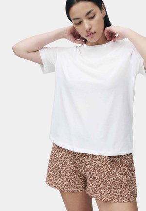 Vrouw die haar haar rechttrekt, gekleed in een effen wit T-shirt met korte mouwen en bruine korte broek met luipaardprint, tegen een lichte achtergrond.