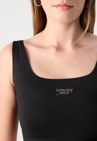 Juodas be rankovių marškinėlis su kvadratiniu iškirpimu, pagamintas iš raštuotos medžiagos. Priekyje, centre, yra sidabrinė siuvinėta "tommy jeans" emblemėlė.