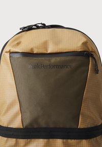 Backpack featuring a tan geometrical grid pattern, olive green panel, black zipper accents, and the "PeakPerformance" logo prominently displayed.