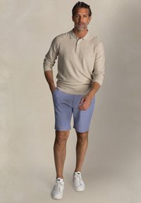 Beige polo shirt met lange mouwen en een gestructureerde brei, gecombineerd met lichtblauwe shorts met een subtiel patroon. Witte sneakers maken de look compleet.