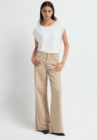 Vrouw in een witte t-shirt met korte mouwen en beige hoge taille wijde broek met kleine stippenversieringen, staande met handen op de heupen.