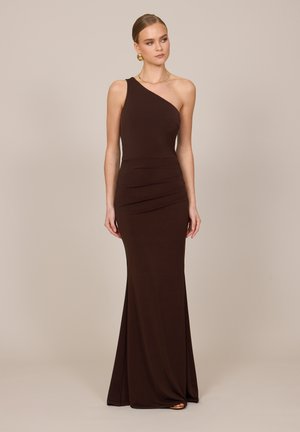 Femme portant une robe longue asymétrique marron foncé avec des fronces subtiles, des boucles d'oreilles anneaux dorées et un fin collier en or, debout sur un fond beige.