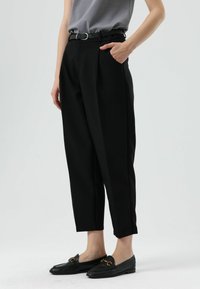 Pantalon noir sur mesure avec plis, doté d'une ceinture noire à boucle argentée. Associé à des mocassins noirs. Tissu lisse, coupe raccourcie.