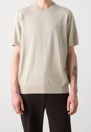 Beiges Stricktop mit kurzen Ärmeln, gerippten Kanten und kleinem "BOSS"-Patch, getragen von einer Person mit tätowiertem linken Unterarm und schwarzen Hosen.